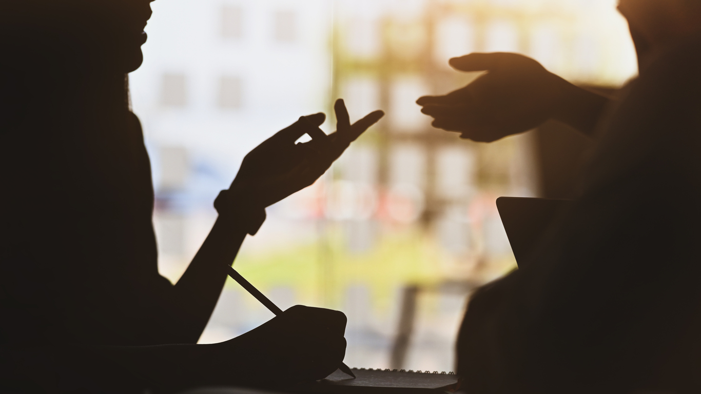 silhouette of young designer team standing with a white blank screen laptop and notebook in hands while discussing/talking about them new project with the modern office as background.